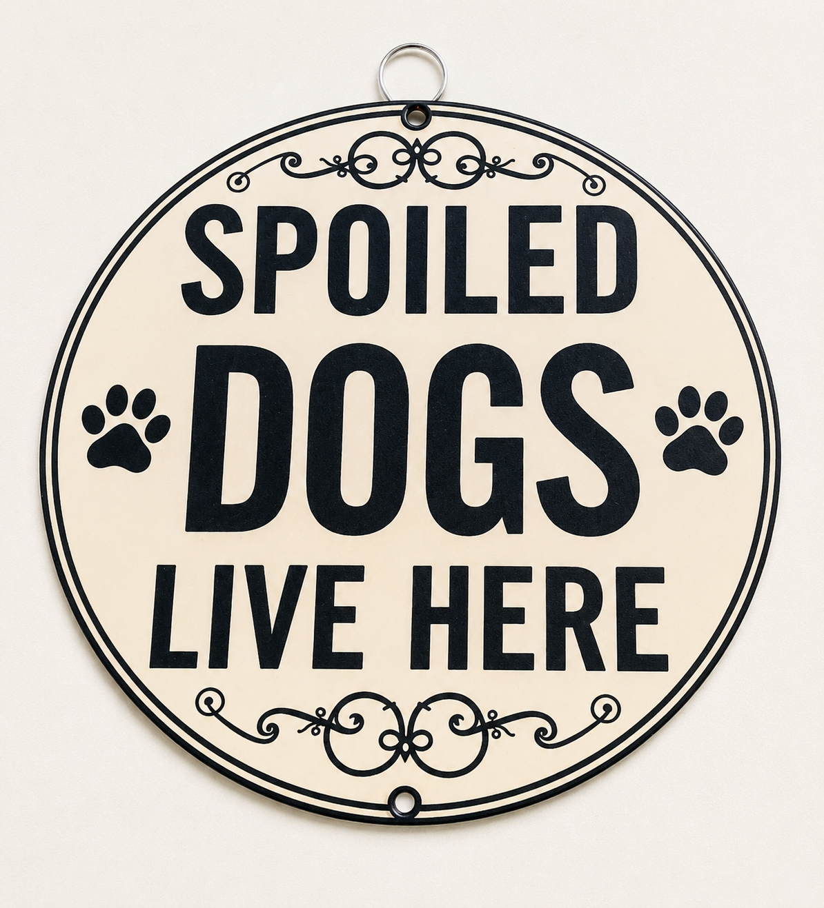 Round decorative wall sign with the phrase 'SPOILED DOGS LIVE HERE' in bold black lettering on a warm cream background, with black paw prints and ornamental scroll details, plus a hanging loop at the top.