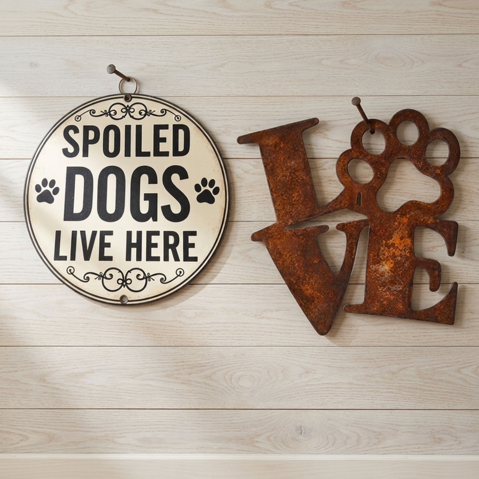 Two rustic metal wall decorations on a light wood plank background: a round cream sign reading 'SPOILED DOGS LIVE HERE' with black text and paw prints, and a rusted brown cutout letter 'LOVE' with the 'O' shaped like a paw print.