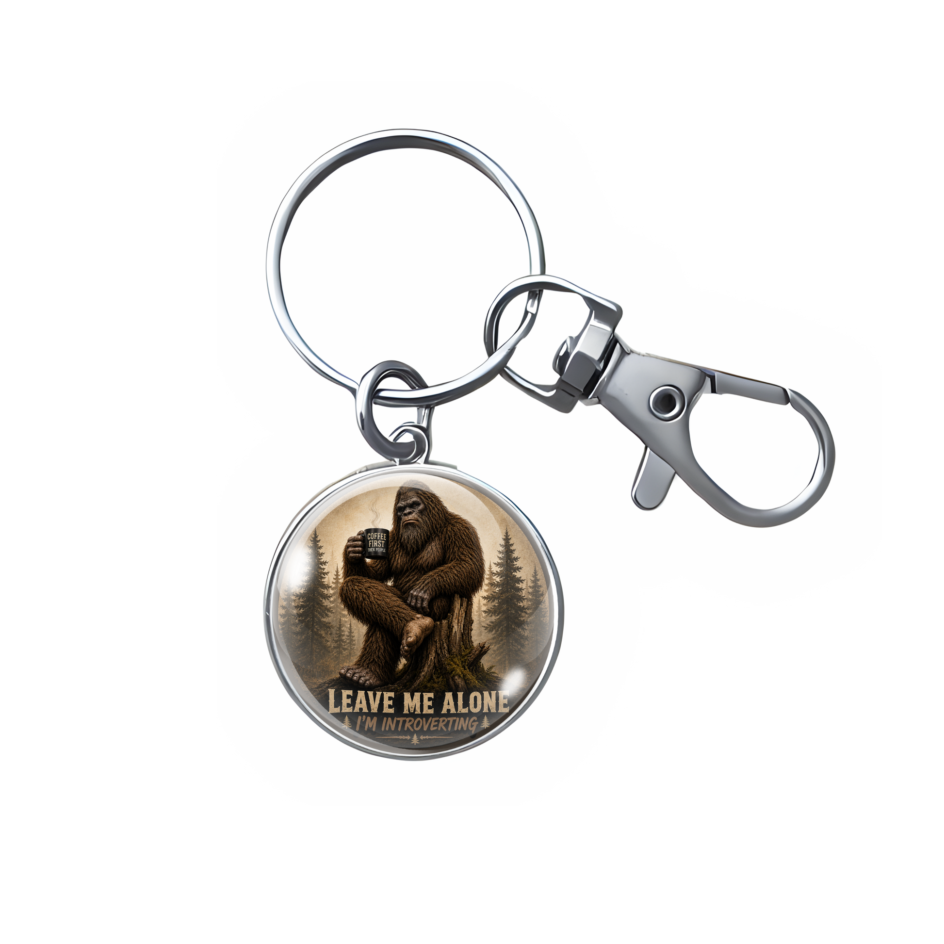 A silver metal keychain with a lobster clasp and split ring, featuring a round charm with an illustration of Bigfoot sitting by a campfire and the text 'LEAVE ME ALONE'.