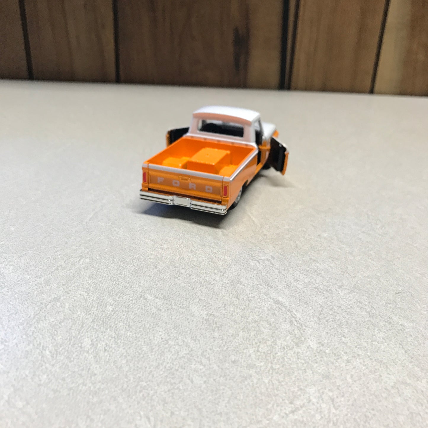 A small orange and white toy dump truck with black wheels, photographed on a light gray textured surface next to a wooden floor.
