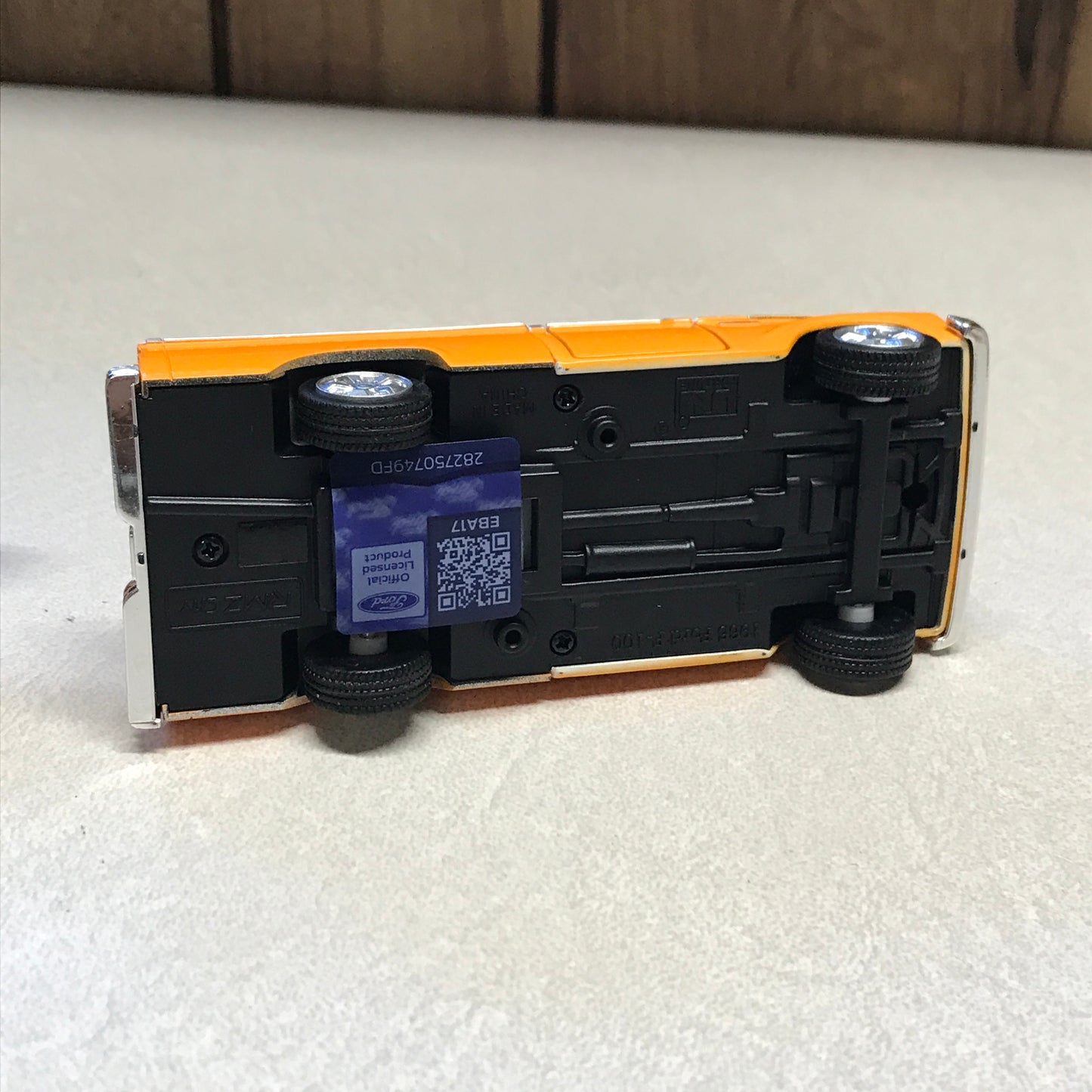 An upside-down toy bus with a black chassis and orange body panels, photographed on a white textured surface with a wooden floor edge visible.