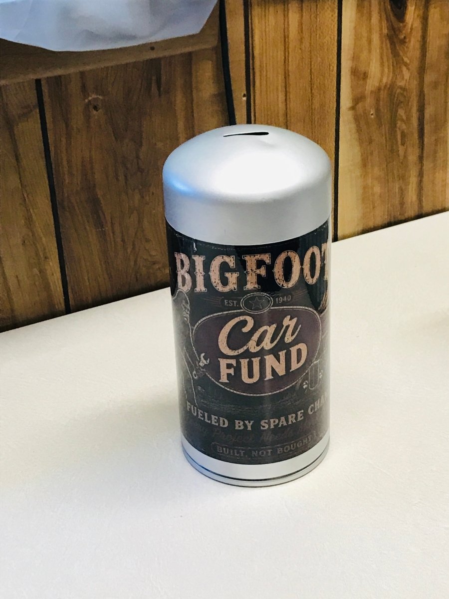 A novelty cylindrical can-shaped container labeled 'BIGFOOT Car FUND' sits on a white tabletop against a wooden paneled background.
