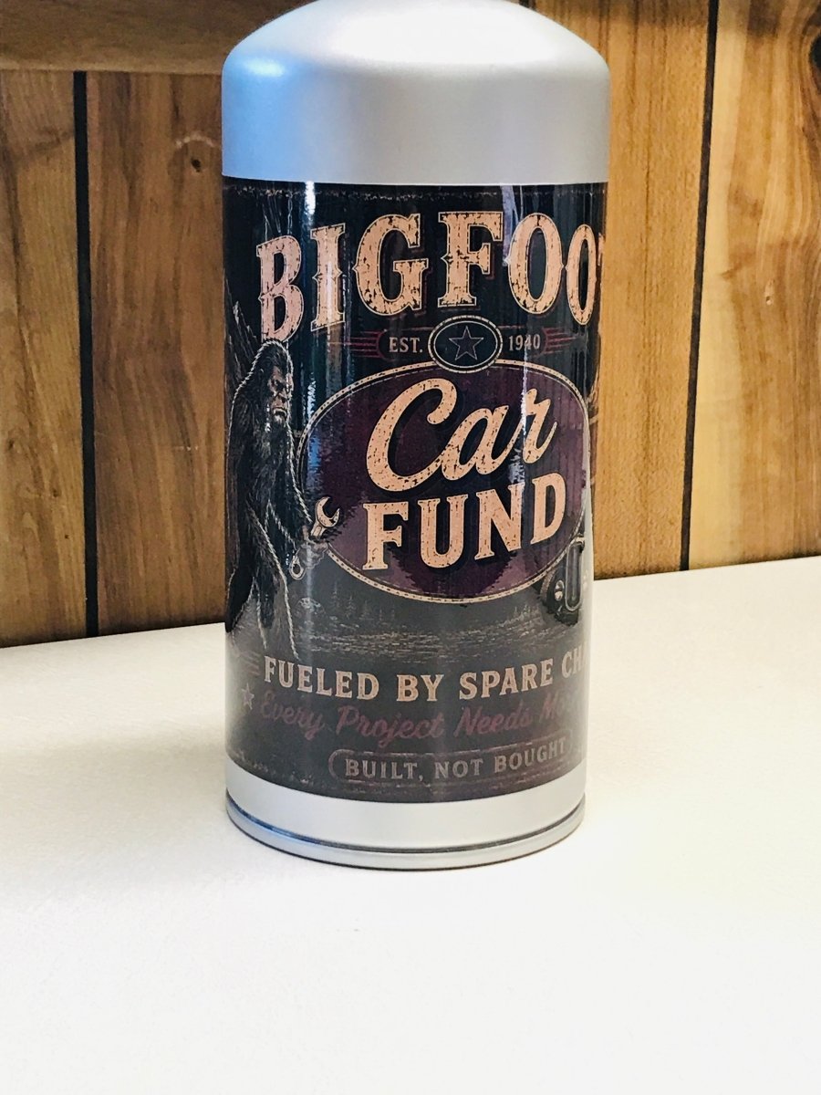 A cylindrical metal can with a white top and bottom, featuring a dark vintage-style label that reads 'BIGFOOT Car FUND' with distressed cream lettering and an illustration of a Bigfoot figure, set against a wood-paneled background.