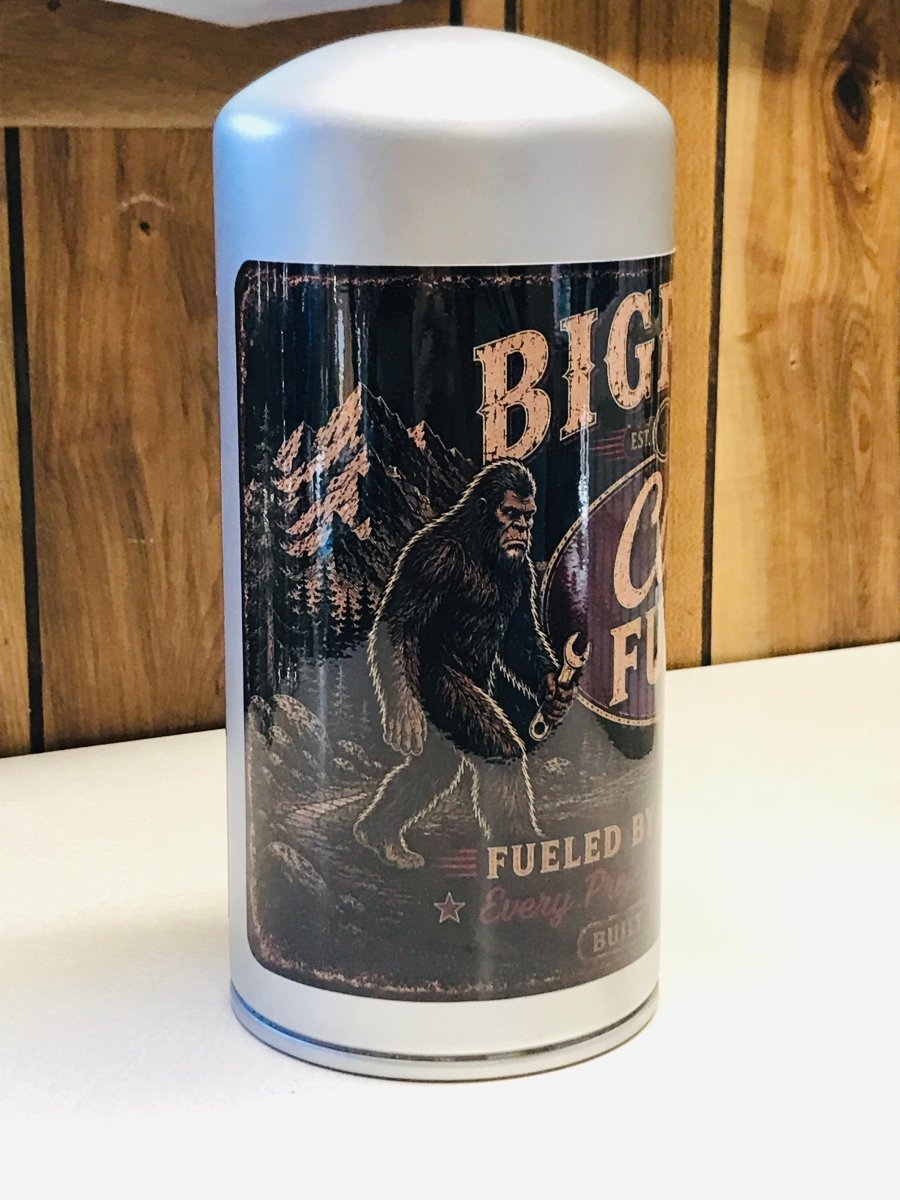 A white cylindrical metal canister with a vintage-style black label featuring a Bigfoot illustration and the text 'BIG FOOT' and 'FUELED BY'. The canister is photographed on a white surface against a wooden background.