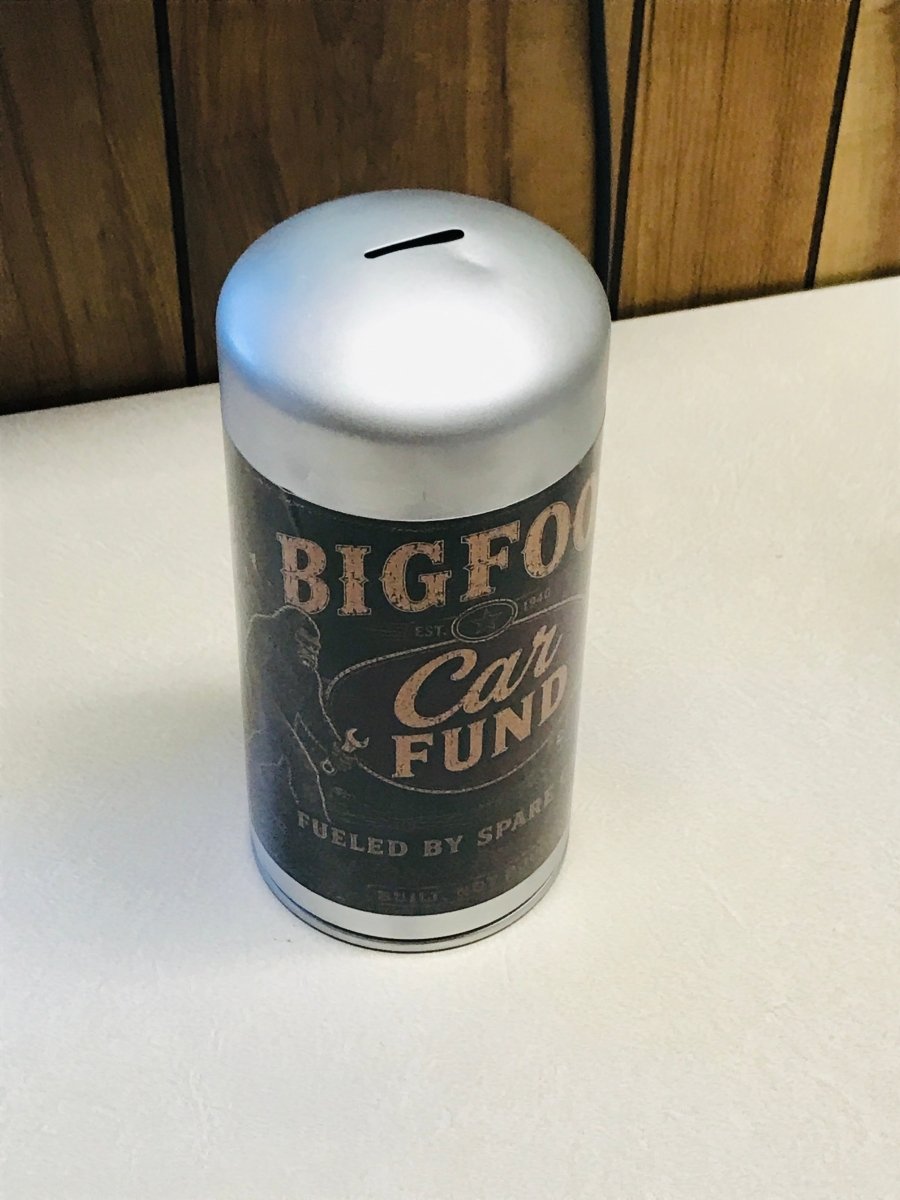 A novelty cylindrical can-shaped pig bank with a silver top and a dark label reading 'BIGFOO Car & FUND' and 'FUELED BY SPARKE', sitting on a light surface against a wood-paneled background.