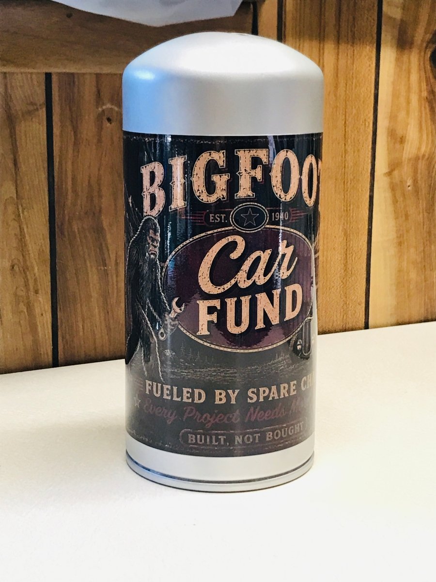 A cylindrical metal canister with a white plastic cap and base, featuring a vintage-style label for 'Bigfoot Car Fund'. The label has a dark background with cream and purple text and graphics.