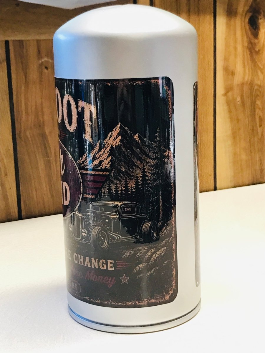 A white cylindrical metal canister with a vintage-style black graphic label featuring a hot rod car, mountains, and distressed text, standing on a white surface against a wood-paneled background.