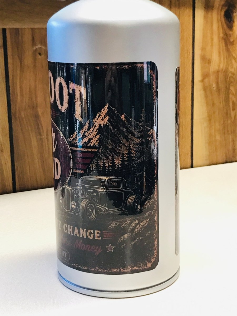 A white cylindrical metal can with a vintage-style black graphic label featuring a hot rod car, mountains, and distressed text, standing on a white surface against a wood-paneled background.