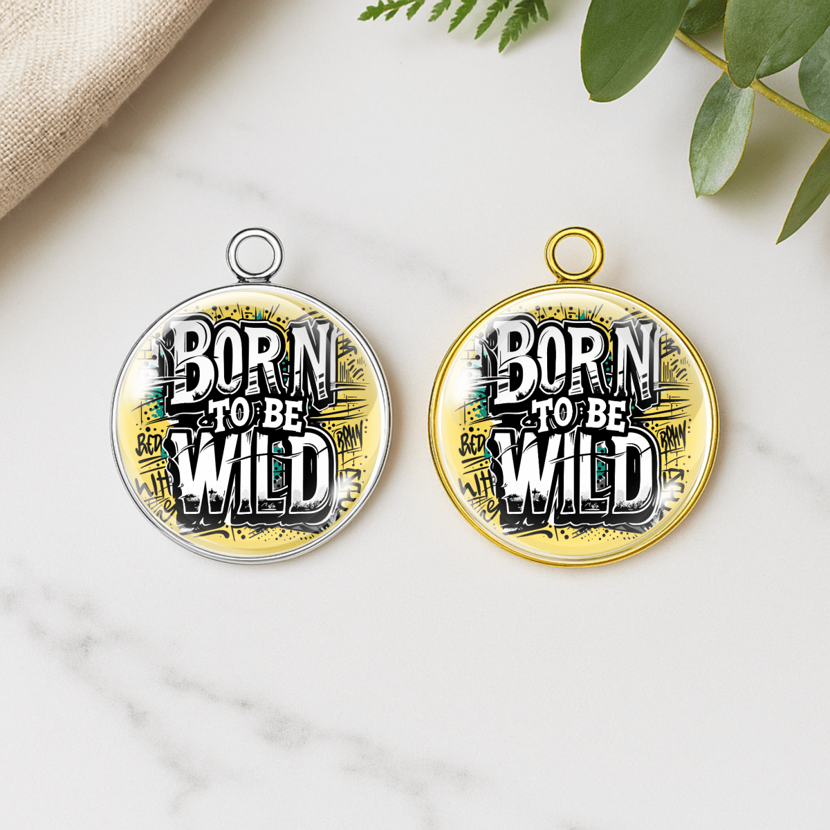 Two round pendants with 'Born to be Wild' text on a marble surface with green leaves.