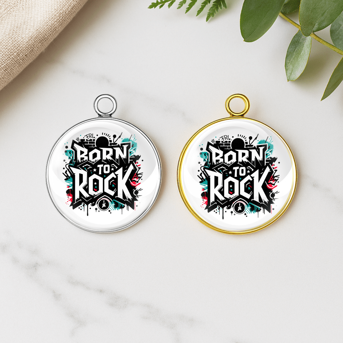 Two 'Born to Rock' pendants on a marble surface with green leaves in the background.
