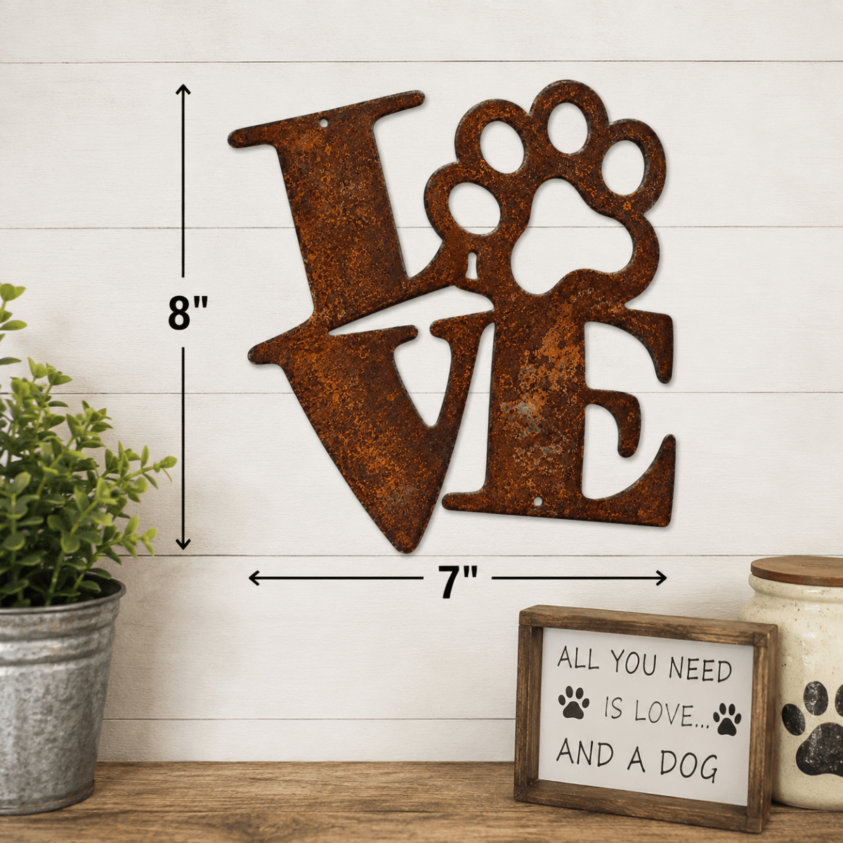 Rustic metal wall decor spelling LOVE with a paw print integrated into the O, shown mounted on a white shiplap wall with dimension arrows indicating 8 inches tall by 7 inches wide. A small potted plant, jar, and sign are visible at the bottom.