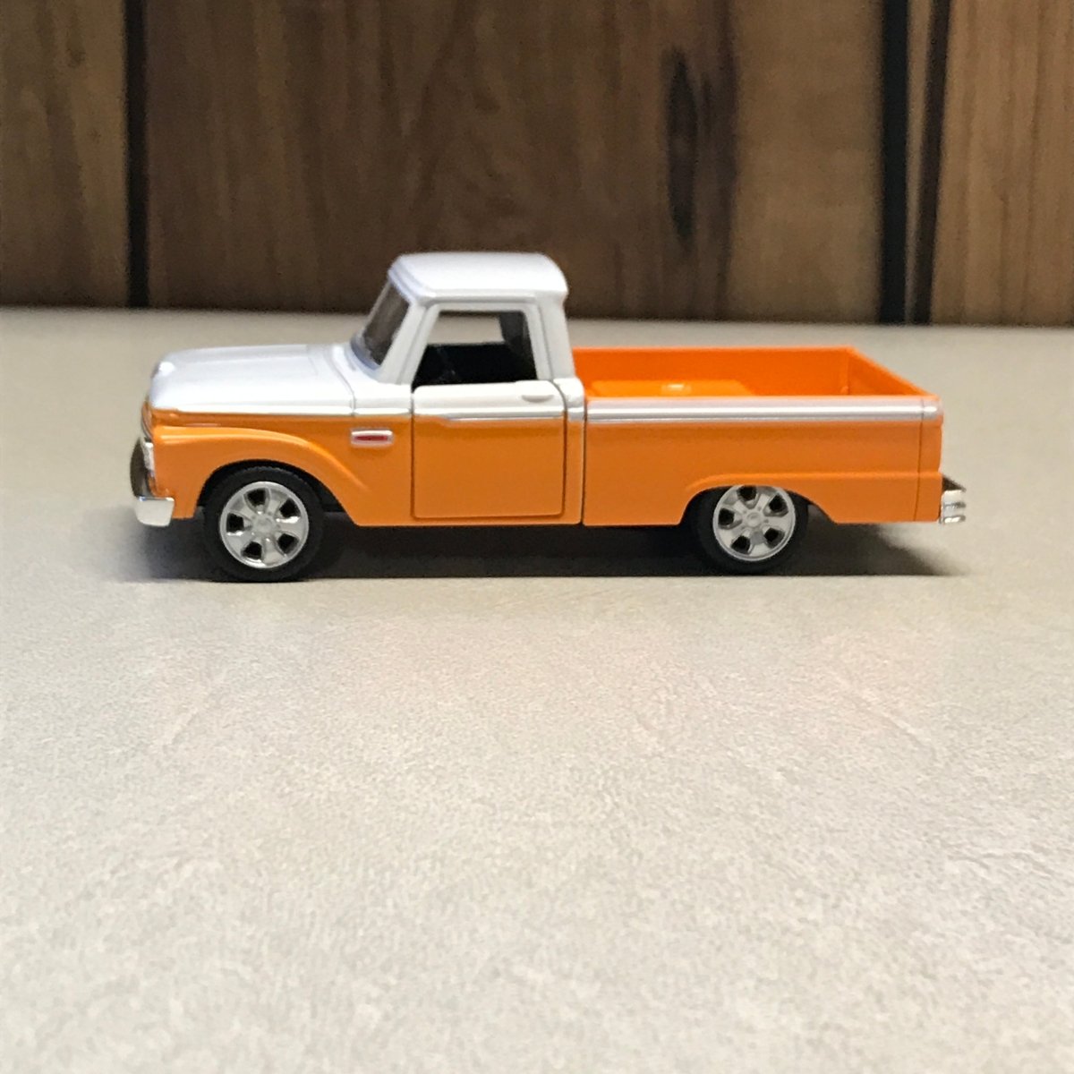 An orange and white toy pickup truck mounted on a textured off-white wall, with a dark wood panel visible on the left side.