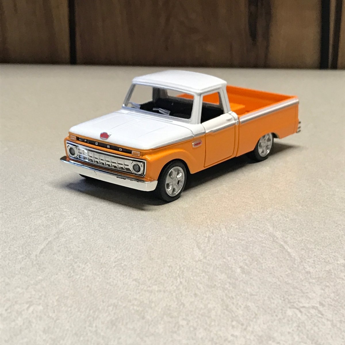 A small orange and white toy pickup truck with black tires, photographed on a light textured surface next to a dark wood area.