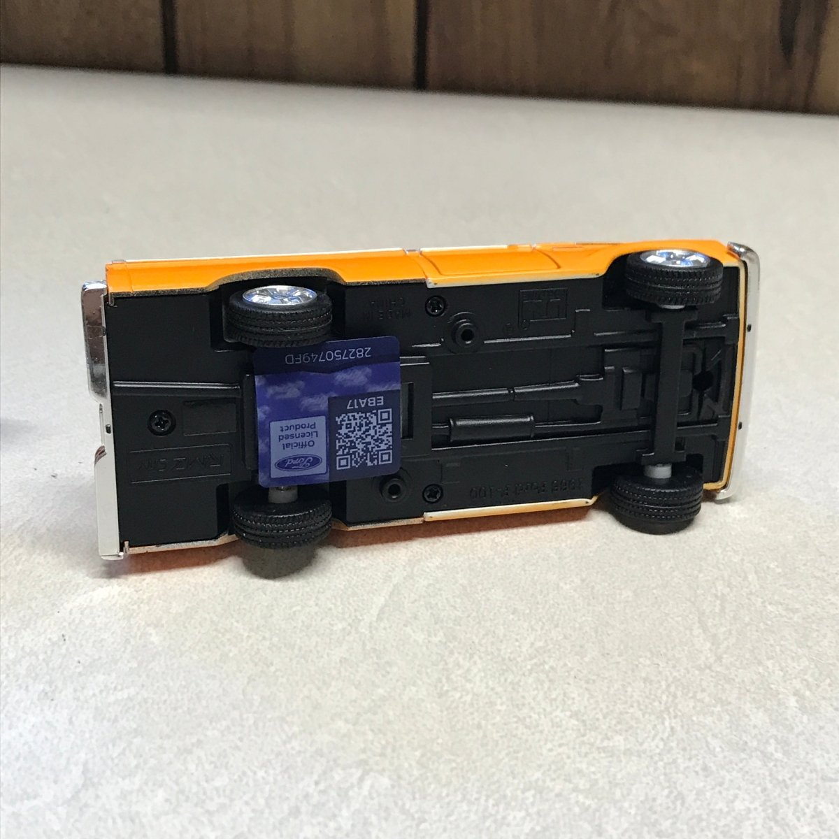An upside-down toy bus with a black chassis and orange body panels, photographed on a white textured surface with a wooden floor edge visible.