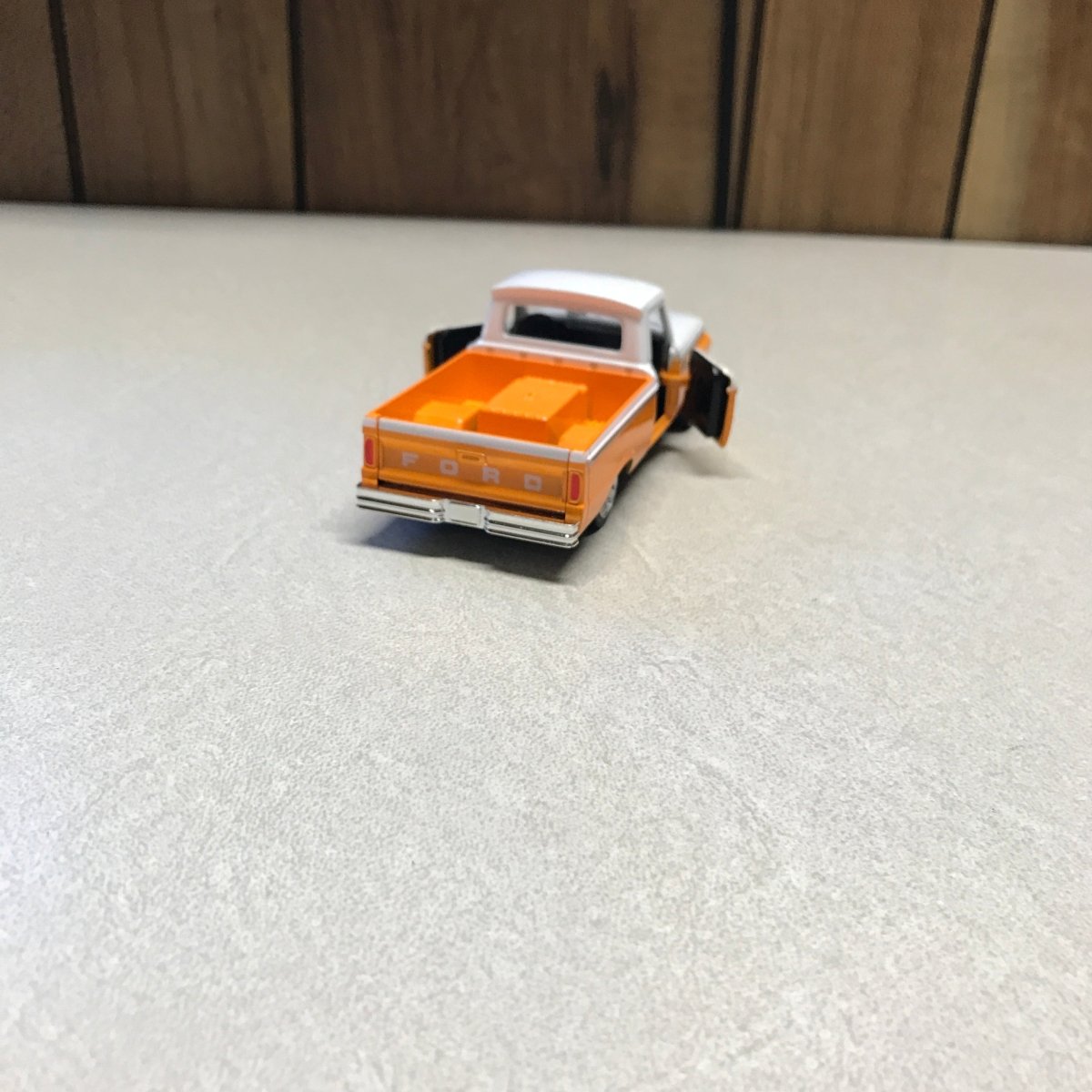 A small orange and white toy dump truck with black wheels, photographed on a light gray textured surface next to a wooden floor.