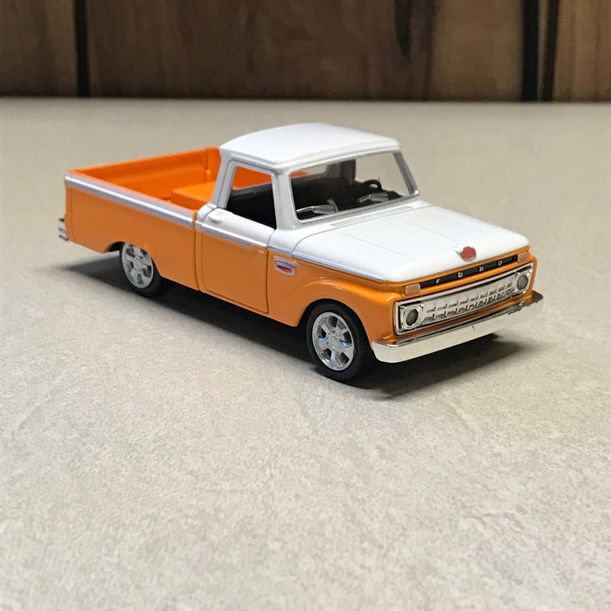A small orange and white toy pickup truck with black tires, photographed on a light textured surface with a dark wood area at the left edge.