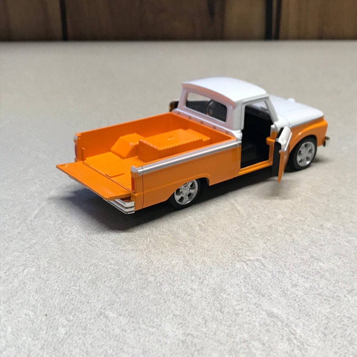 A small orange and white toy dump truck with a black bed, photographed on a light textured surface.