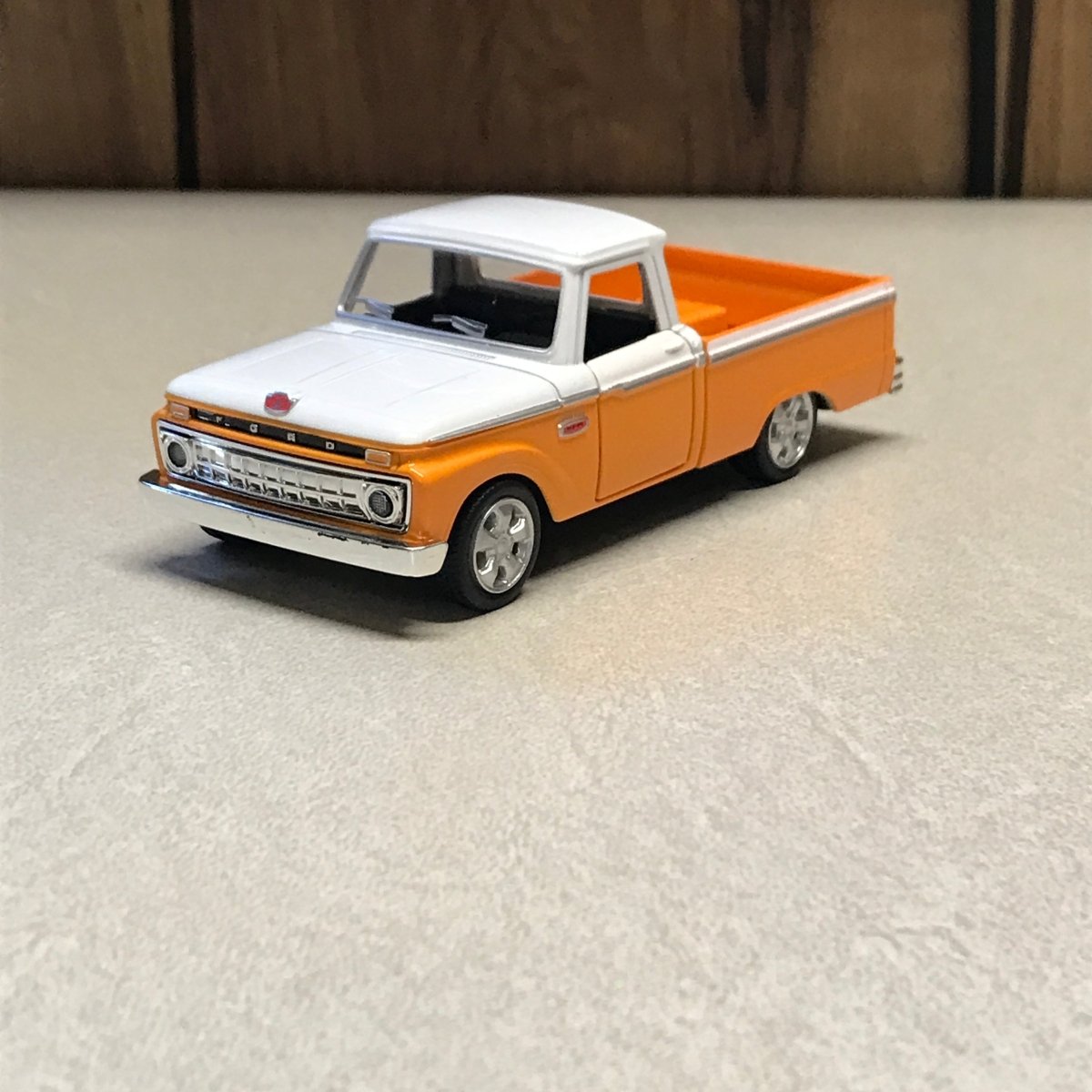 A small orange and white toy pickup truck with black tires, photographed on a light beige textured surface with a dark wood area at the left edge.