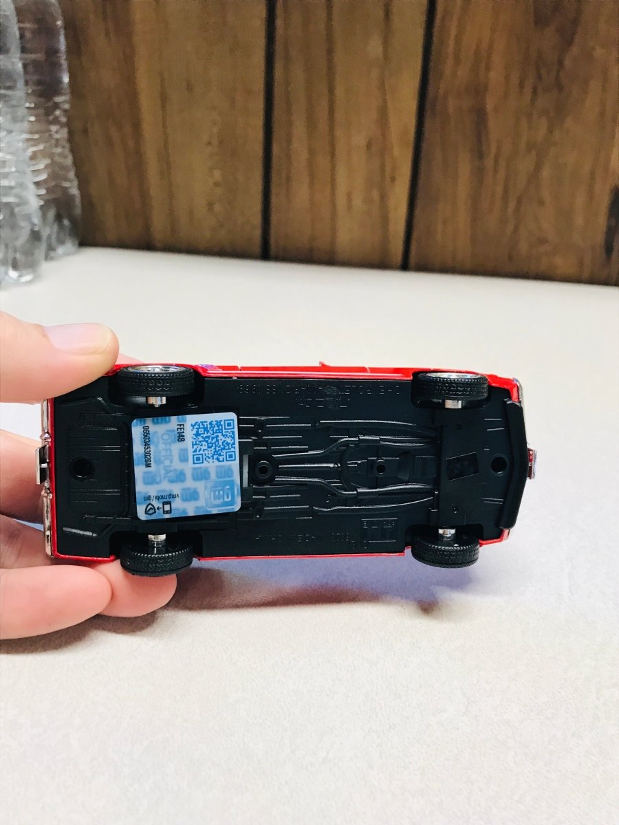 A hand holding a red toy car upside down to show the black plastic undercarriage and a blue sticker with a QR code, set against a white tabletop and wooden background.