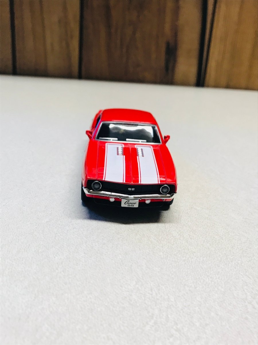 A red die-cast model car with white racing stripes on the hood, positioned on a light gray textured surface with a wooden background.