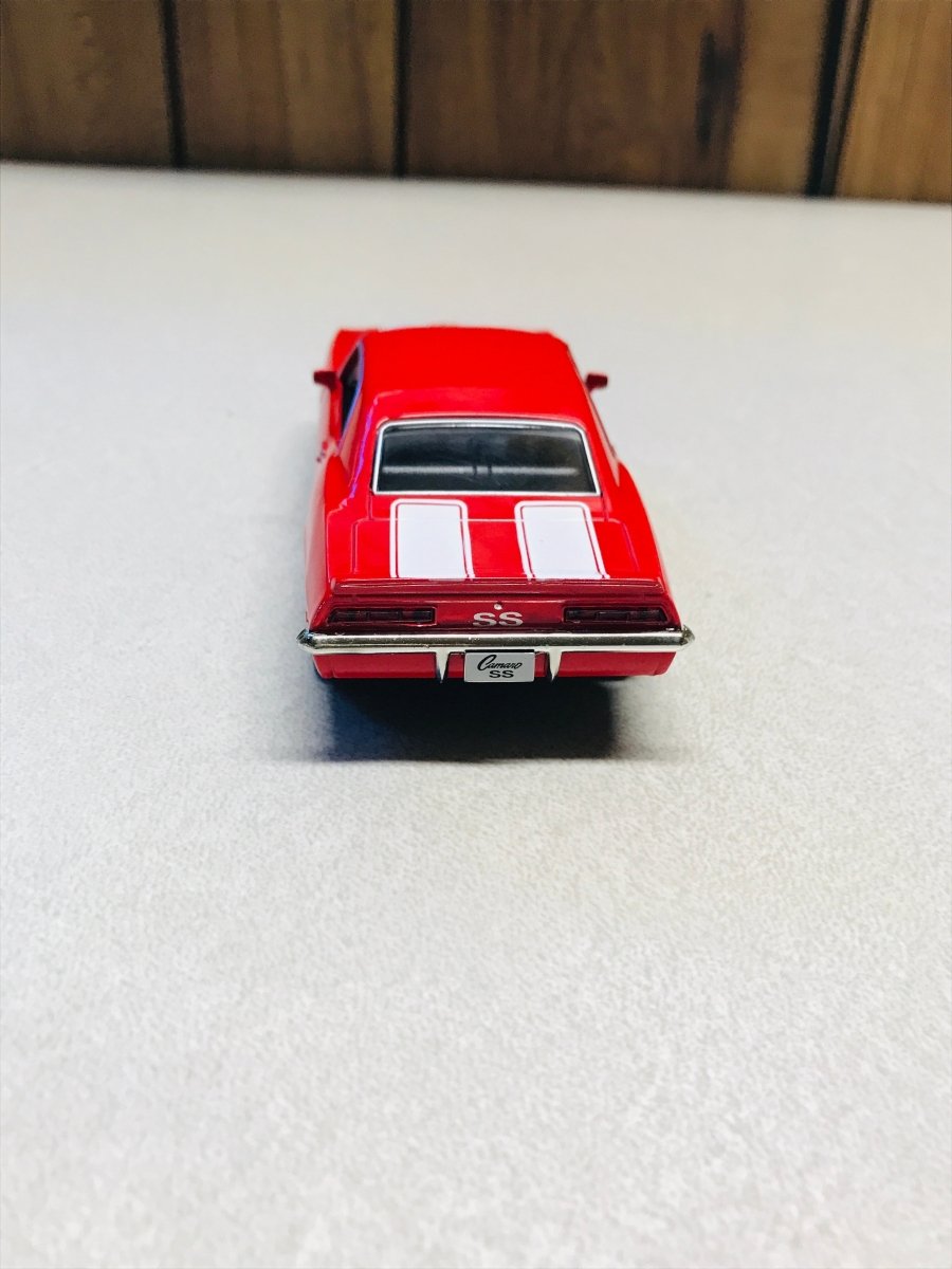 A red die-cast model car with white racing stripes on the roof, viewed from the rear on a light gray surface with a wooden background.