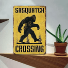 Wooden sign with 'Sasquatch Crossing' design on a wooden surface with a plant in the background