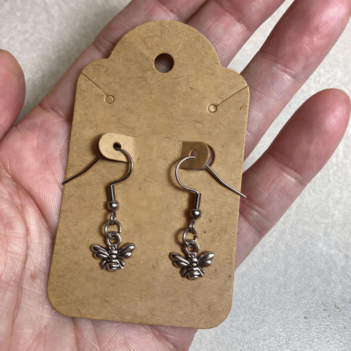 Silver bee-shaped earrings on a brown cardboard backing held in a hand.