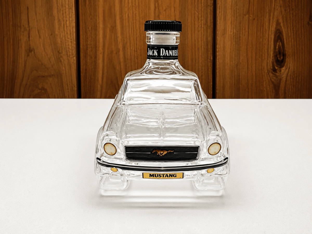 A clear glass bottle shaped like a classic Ford Mustang car, with a black neck label reading and a gold Mustang badge on the front, set on a white surface against a wooden background.