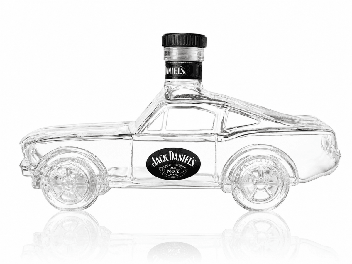 A clear glass bottle shaped like a car, featuring a black cap and a prominent black oval Jack Daniel's logo on the side, set against a white background with a soft reflection below.