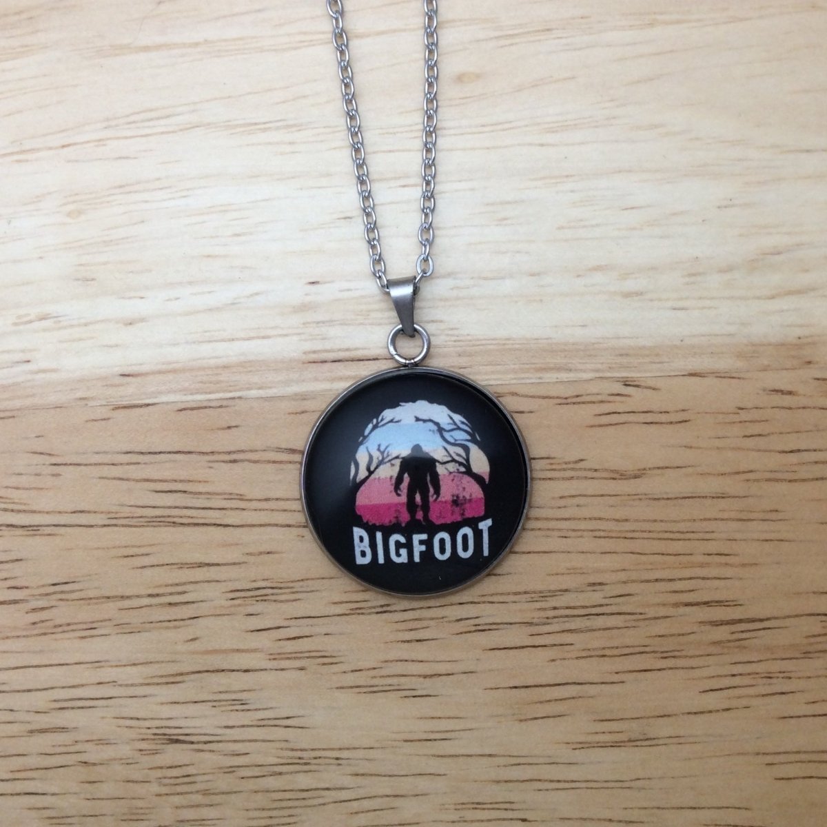 Stainless steel necklace with a glass cabochon pendant that Showa a Bigfoot walking in front of a colorful moon and it says "Bigfoot"