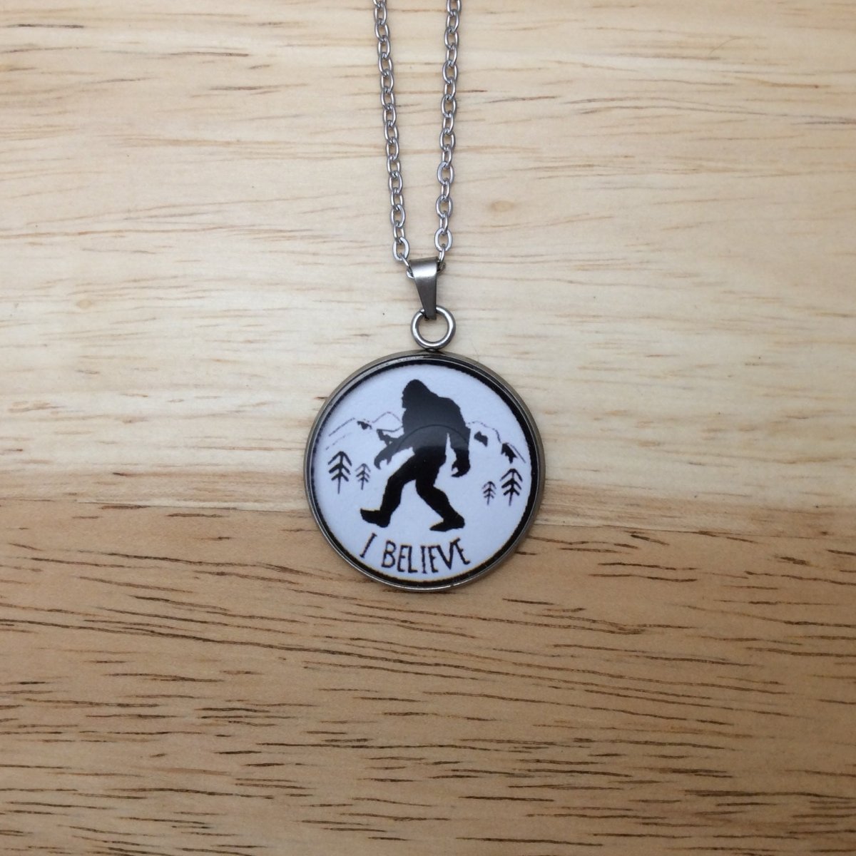 Stainless steel necklace with a glass cabochon pendant that Showa a Bigfoot walking in front of mountains and it says "I Believe"