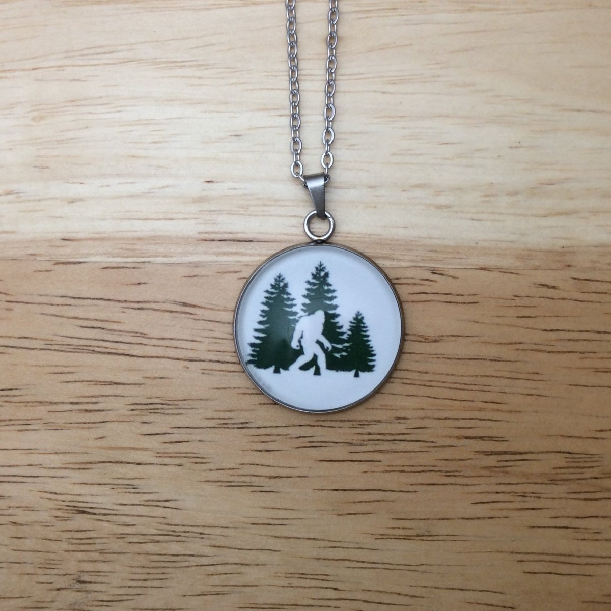 Stainless steel necklace with a glass cabochon pendant that Showa a Bigfoot walking in front of  Green Pine Trees