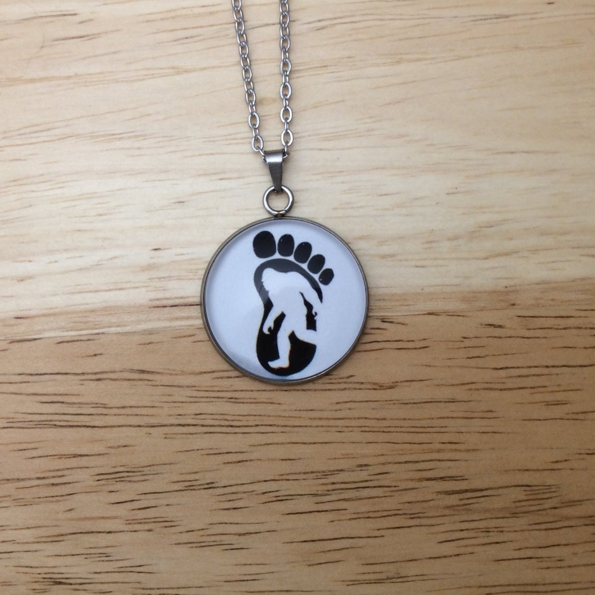 Stainless steel necklace with a glass cabochon pendant that Showa a Bigfoot walking in  an image of a big foot.