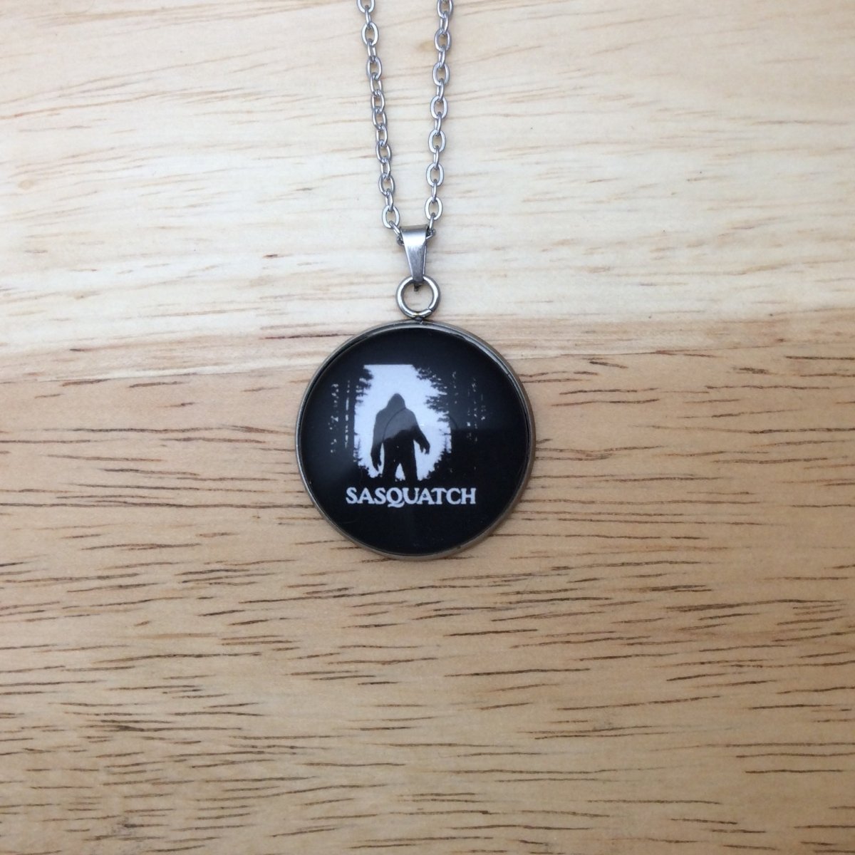 Stainless steel necklace with a glass cabochon pendant that Showa a Bigfoot walking through the forest in front of a white moon and it says "Sasquatch"