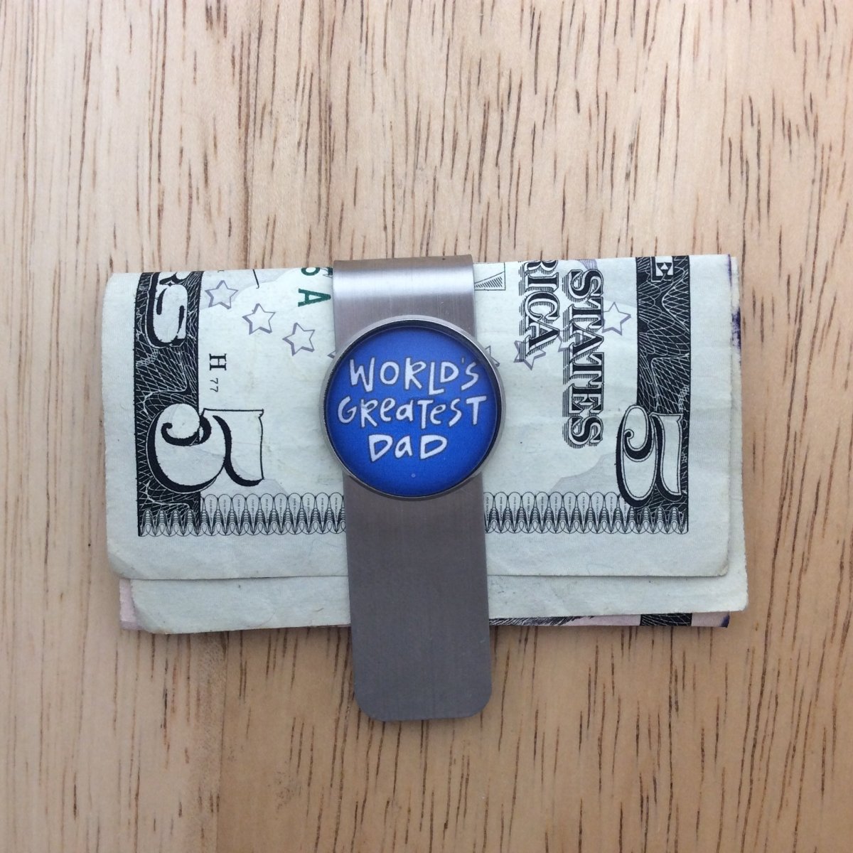 stainless steel glass cabochon money clip holding a five dollar bill. The cabochon is blue with whit lettering that says "worlds Greatest Dad"